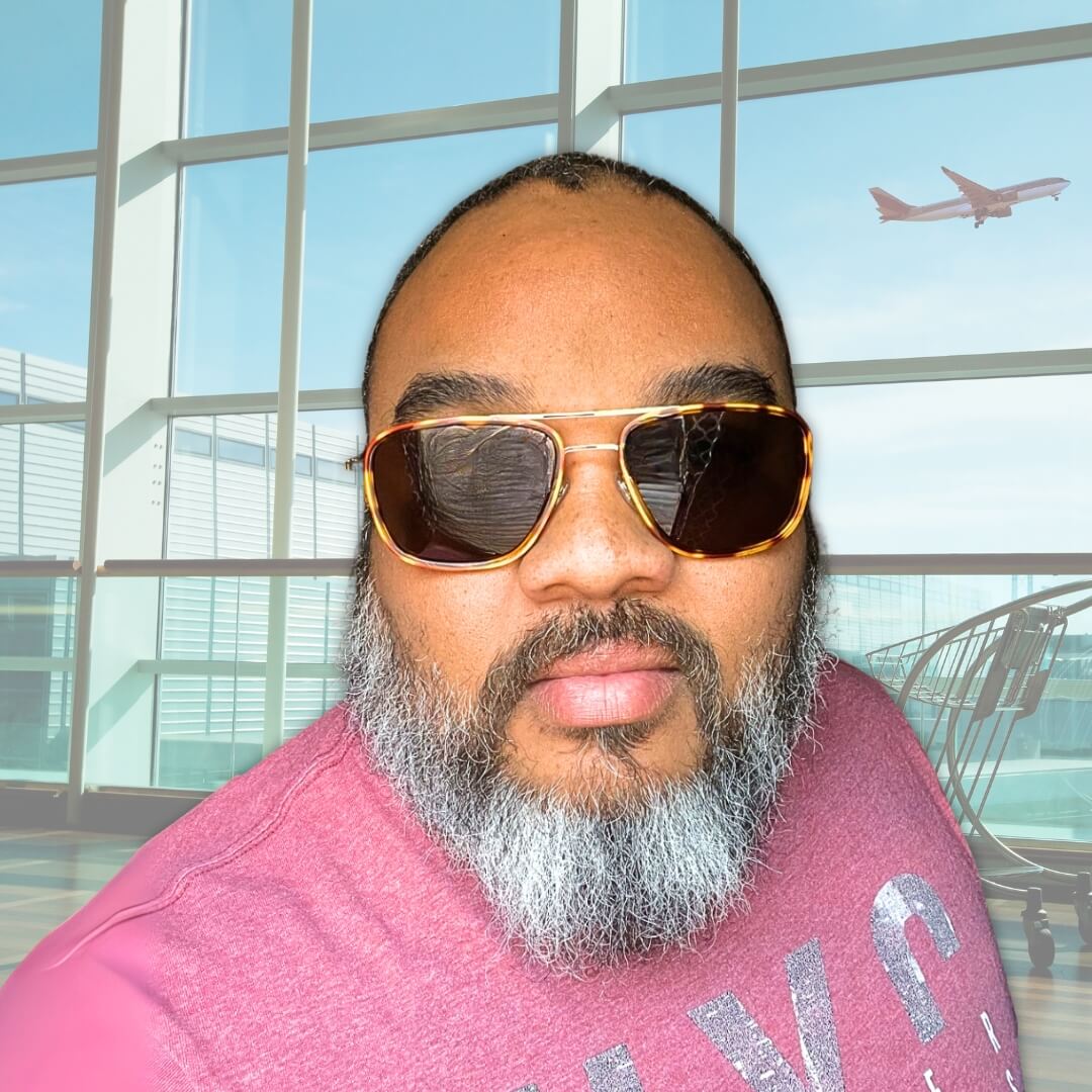 Confident man with a salt-and-pepper beard wearing tortoise-shell big sunglasses for big heads with amber lenses, standing in front of modern glass windows and an airplane in the background.