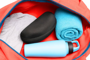 Extra large black sunglasses case inside a red gym bag, shown alongside a folded towel, gray t-shirt, and light blue water bottle.