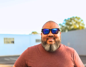 Bearded man wearing Faded Days 165mm XXL sunglasses with blue mirrored lenses, smiling outdoors in a red shirt.