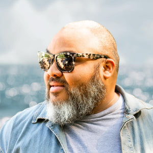 Man wearing tortoiseshell Robust big head sunglasses, looking left with ocean behind him