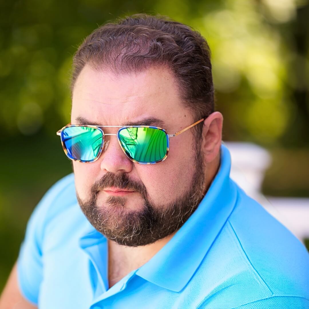 Serious-looking man in a blue polo shirt wearing mirrored big sunglasses for big heads, seated outdoors with a blurred green background.