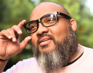 Bearded man wearing wide-fit Faded Days prescription glasses with clear lenses, looking up while adjusting frame outdoors.