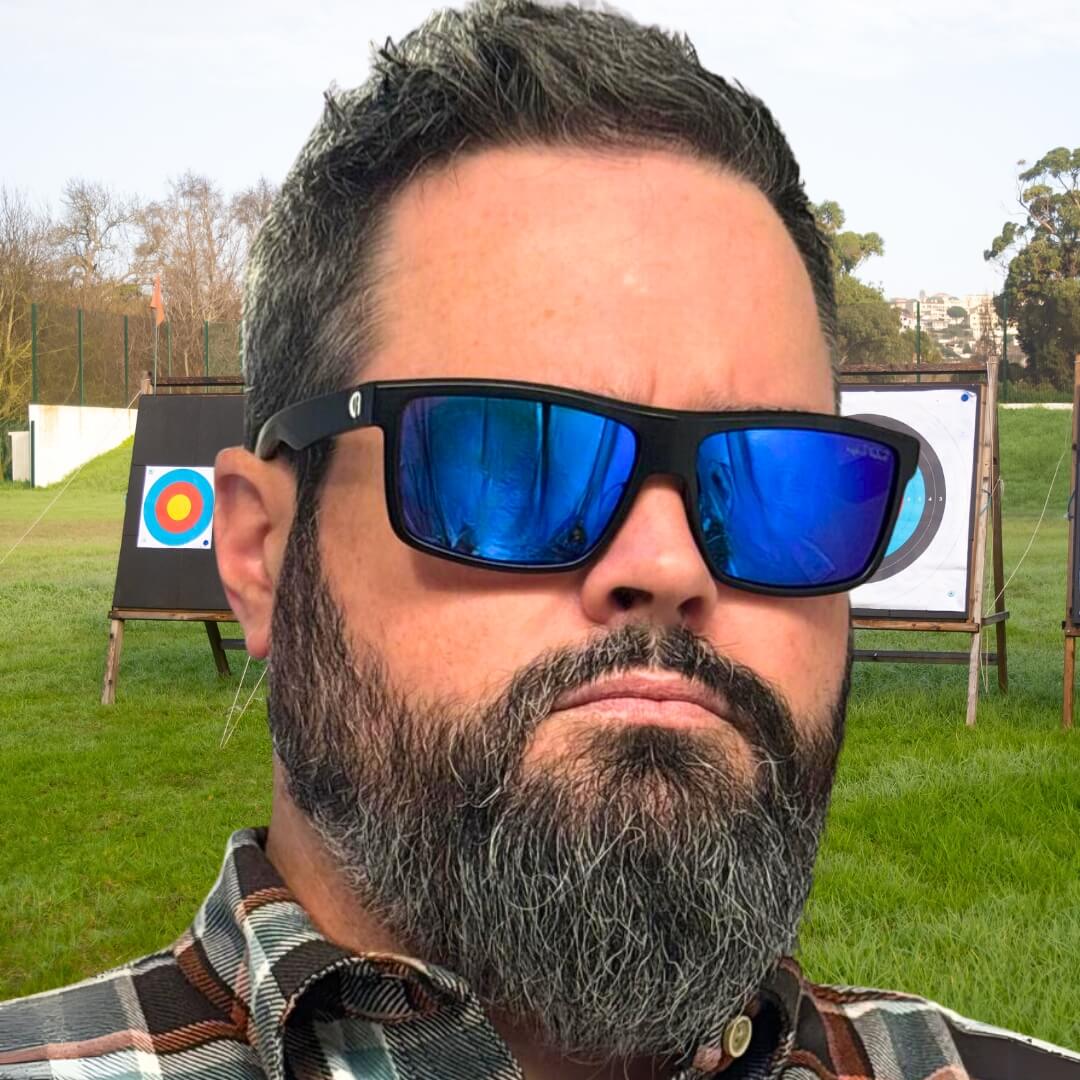 Man with dark hair and beard wearing mirrored blue-lens Faded Days sunglasses for big heads, standing near an archery target.